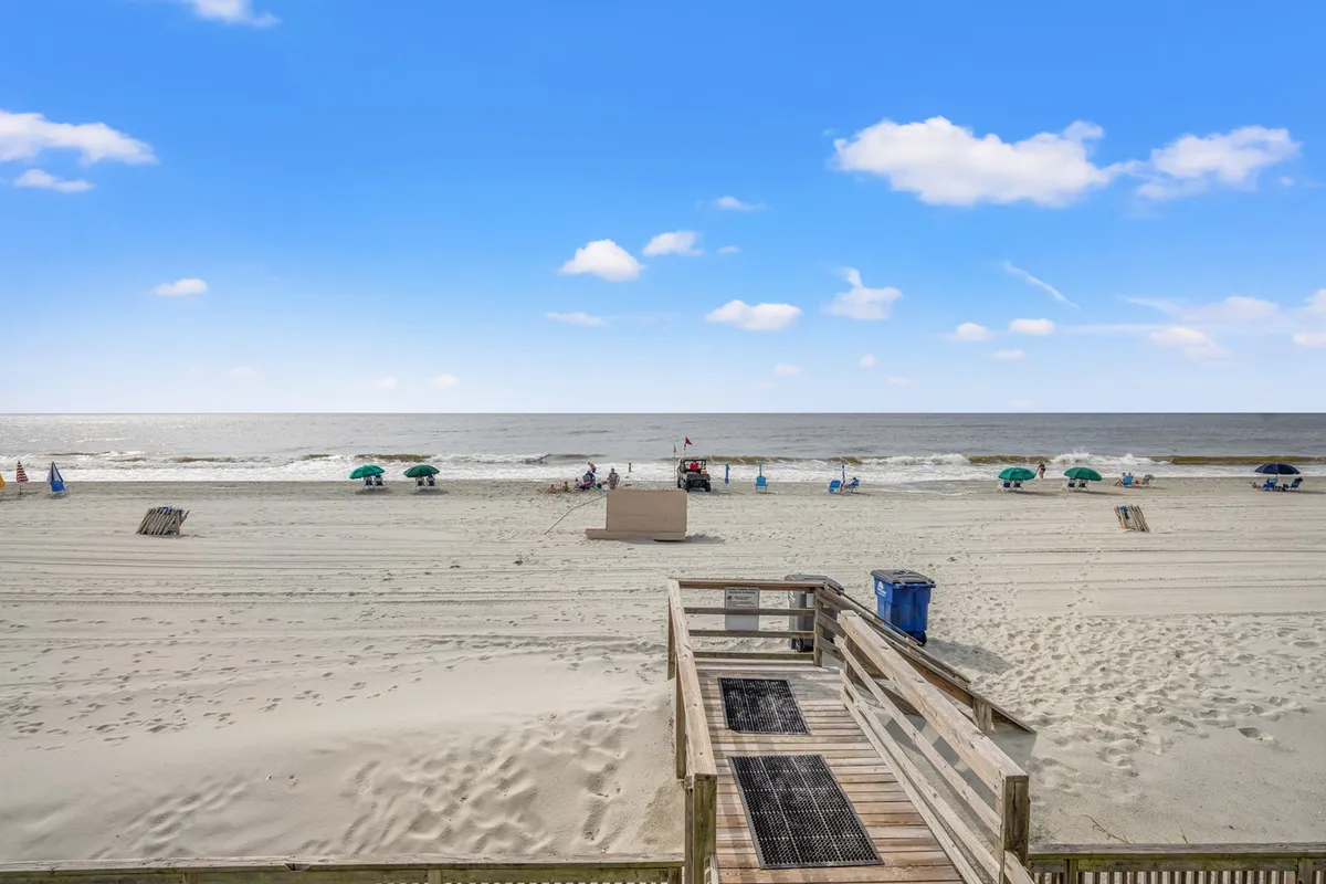 A scenic beachfront view featuring the ocean, umbrellas, people sunbathing, and a boardwalk, evoking the sense of private beach access and leisurely coastal living. The scene is lively yet peaceful, capturing the unique draw of the property's location.