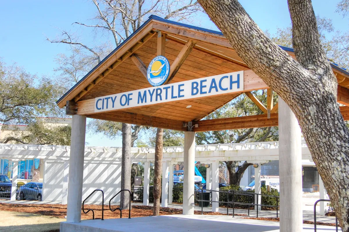 A welcoming exterior scene featuring a wooden structure with the 'CITY OF MYRTLE BEACH' sign, surrounded by lush trees. The space suggests a community gathering area, ideal for local events, outdoor enjoyment, and social meetups.