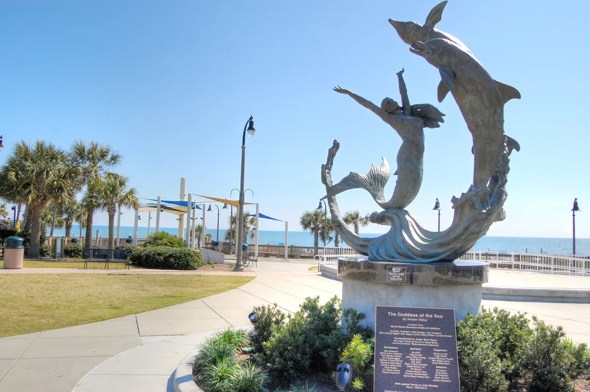 A scenic coastal setting featuring the iconic sculpture of the Goddess of the Sea, with palm trees and a wide view of the ocean. People are seen strolling, taking photos, and relaxing in the sunshine.