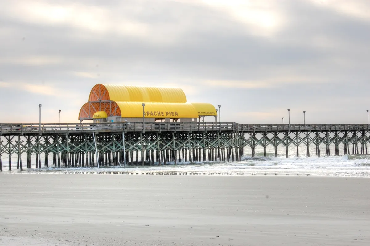 This image showcases the iconic Apache Pier, with its distinctive yellow roof extending over the shimmering ocean. Waves crash against the pier's base, while a tranquil atmosphere invites leisurely strolls and fishing. The wide view allows for a sense of openness and the beauty of Myrtle Beach's oceanfront.