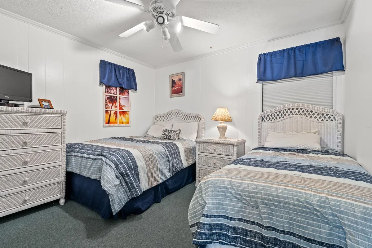 This image shows a tranquil bedroom with two beds featuring striped bedding, a nightstand with a lamp, and a dresser topped with a TV. The space is softly lit, evoking a feeling of relaxation and comfort—perfect for families or groups sharing a room. Sunlight streams in from the window, and the arrangement highlights the flexibility of sleeping options.