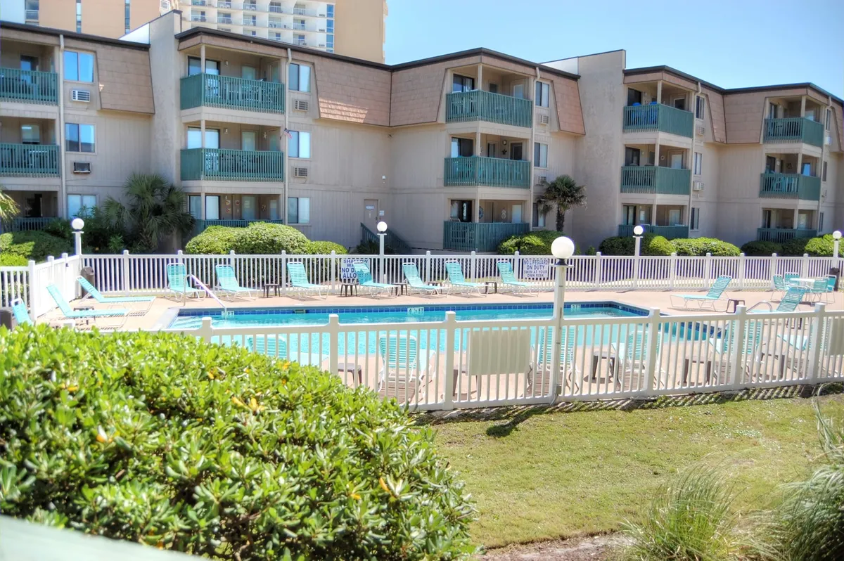 A sparkling resort-style swimming pool is surrounded by lounge chairs, lush landscaping, and overlooks the balconies of vacation condos. Families relax by the water, bask in the sun, or enjoy a morning swim.