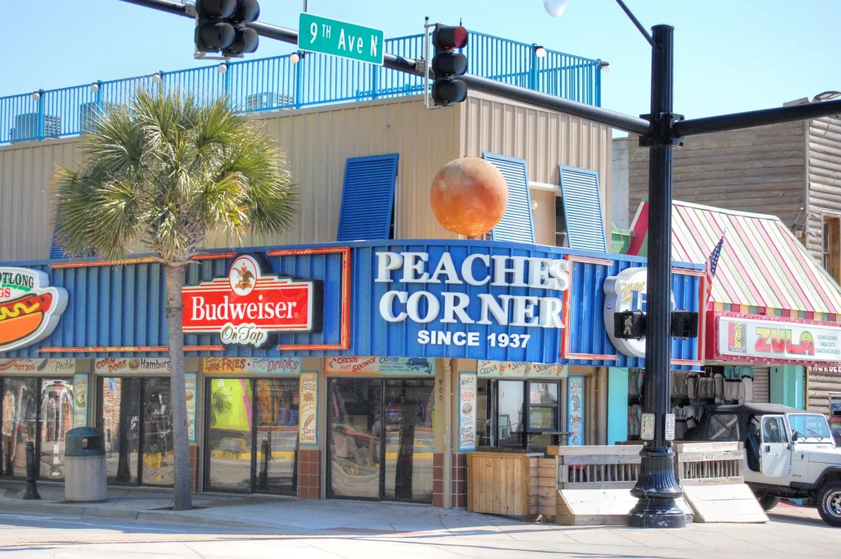 This vibrant exterior shot shows the iconic Peaches Corner sign, a Budweiser sign, and several colorful building facades, with groups of people dining, shopping, and socializing outside.
