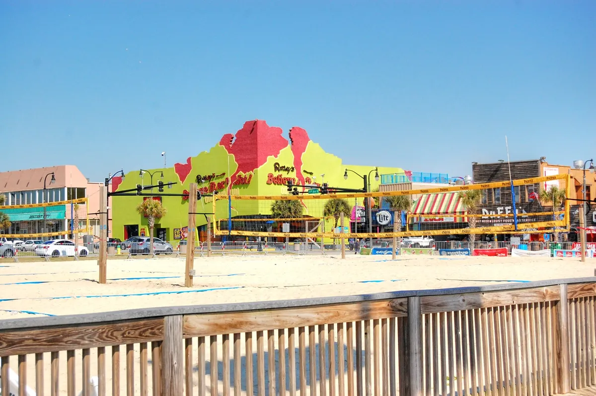 The image captures a lively stretch of Myrtle Beach: a colorful building facade, beach volleyball courts, and a wooden boardwalk bustling with activity. The scene is vibrant yet approachable, reflecting opportunities for casual dining, local shopping, and beachside fun.