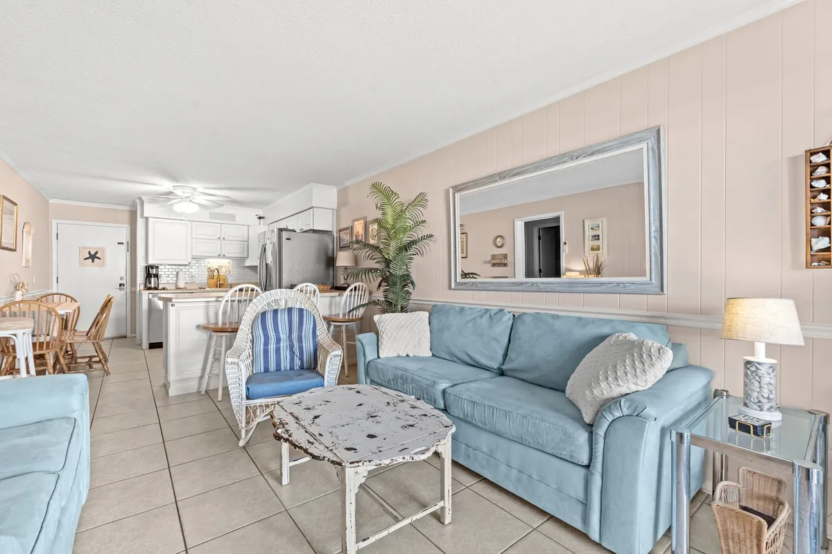 A cozy, open-concept living and dining area featuring blue sofas arranged for conversation, a wooden dining set perfect for gatherings, a unique coffee table, and a decorative mirror with a potted plant adding a touch of greenery. The space is bathed in natural light, accentuating the inviting, family-friendly atmosphere.