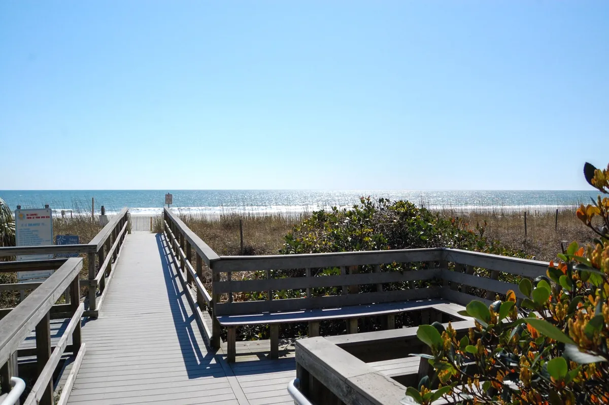 A serene exterior scene with a wooden boardwalk leading through dune grass towards the ocean, offering expansive water views and a tranquil beachside vibe. The scene evokes exploration, relaxation, and the beauty of the Myrtle Beach setting.