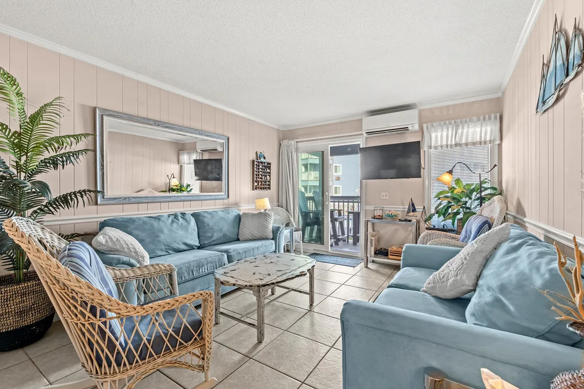 A bright, casual living room with light blue sofas, a decorative mirror, tropical plant, and sliding glass doors that open to a balcony. The space is filled with natural light, providing a warm setting for relaxing, entertaining, or enjoying the view.