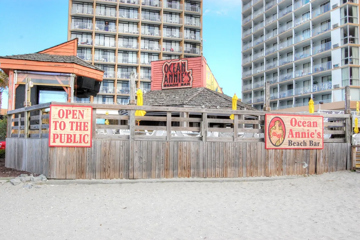 This image features the Ocean Annie's Beach Bar signage, a welcoming wooden deck, and beach chairs under umbrellas, all set against the backdrop of the ocean. The scene illustrates people socializing, enjoying drinks, and listening to live music in a breezy, open-air seaside setting.