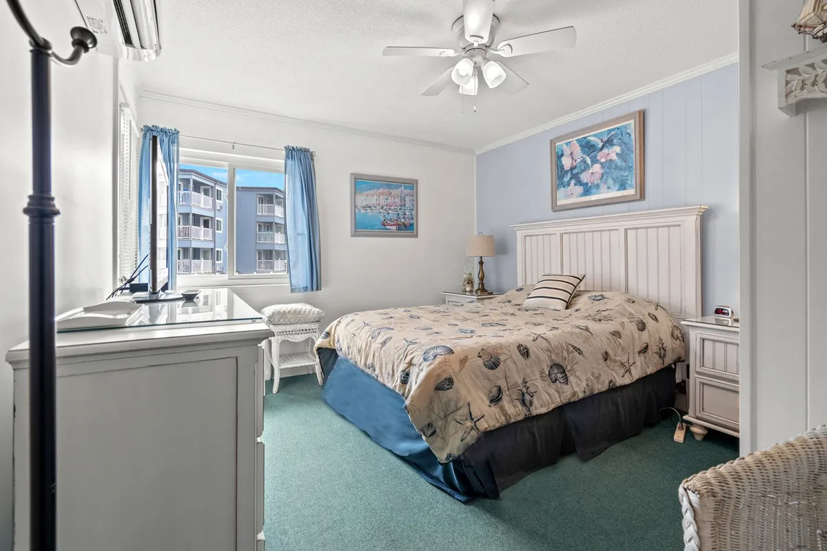 A serene bedroom is depicted with a cozy bed covered in a decorative quilt, a large window adorned with blue curtains, and framed artwork on the walls. The soft natural light enhances the soothing ambiance, evoking the calming coastal décor described in the content.