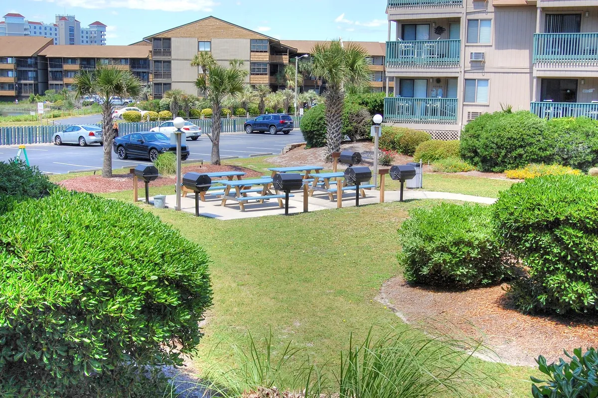 An outdoor grilling area complete with picnic tables, landscaped greenery, and surrounding residential buildings. The space is set up for casual barbecues and group gatherings, emphasizing the communal and social aspects of the property.