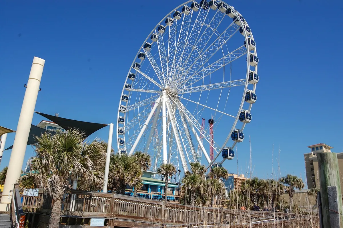 The image features the iconic Myrtle Beach SkyWheel towering above the boardwalk, flanked by palm trees and set against a clear blue sky. Visitors stroll along the boardwalk, enjoying the vibrant beach atmosphere and scenic views of the ocean.