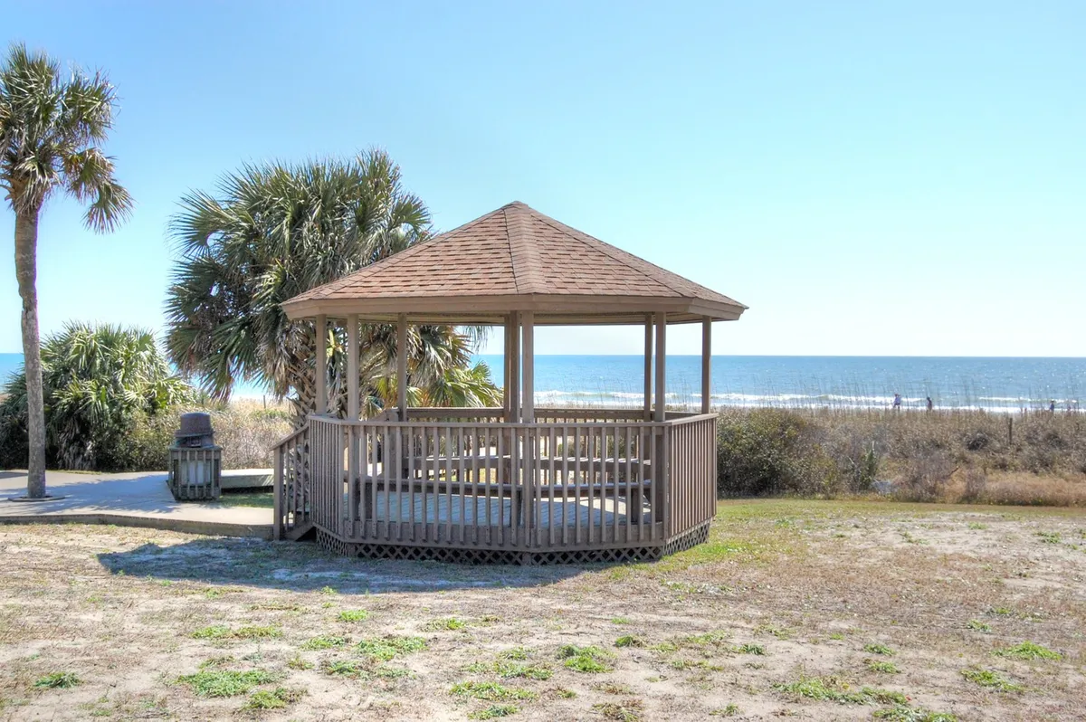 A tranquil wooden gazebo sits on the edge of the sandy beach, framed by palm trees and looking out onto the ocean. The inviting setting is ideal for sunbathing, picnicking, or gathering with family and friends for a shaded break by the sea.
